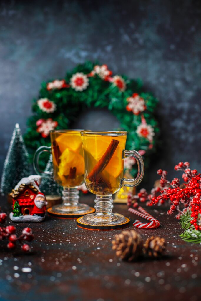 Two glasses of festive spiced Christmas punch garnished with cinnamon sticks, set against a holiday backdrop with a wreath, red berries, and decorative ornaments.