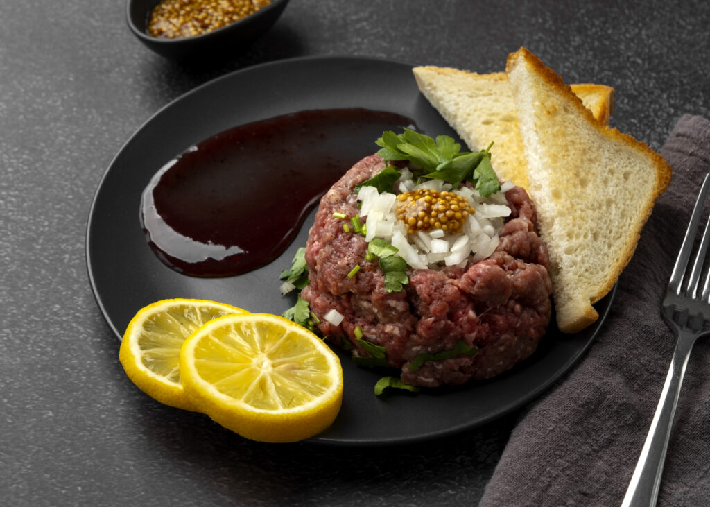 Tartaar in de airfryer served as a gourmet dish with parsley, onions, mustard seeds, lemon slices, and toasted bread on a black plate.